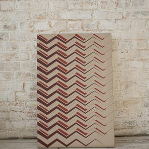 Könnte beinhalten: Ein beigefarbener Leinwanddruck mit einem geometrischen Muster aus roten und braunen Zickzacklinien.