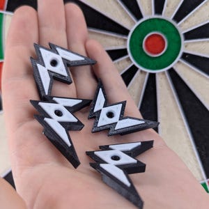 Op de afbeelding: Een verzameling zwarte en witte dartpijlvleugels in de vorm van bliksemschichten. De vleugels worden in een hand gehouden, met een dartbord op de achtergrond. Het dartbord heeft een groen, rood, zwart en wit kleurenschema.
