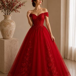 May include: A floor-length, off-the-shoulder ball gown in deep red. The bodice features intricate lace detailing and a sweetheart neckline. The skirt is crafted from layers of tulle, accented with vertical floral lace appliqués that extend from the waist down to the hemline.