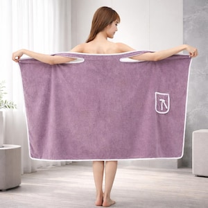 May include: A lavender-colored wrap towel with white trim and shoulder straps. The towel has a pocket with a white embroidered design. The person is holding the towel open, showcasing its full size and design.
