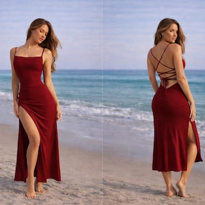 May include: A burgundy-colored maxi dress with a high slit and spaghetti straps. The dress features a crisscross back design and is worn on a sandy beach with the ocean in the background.