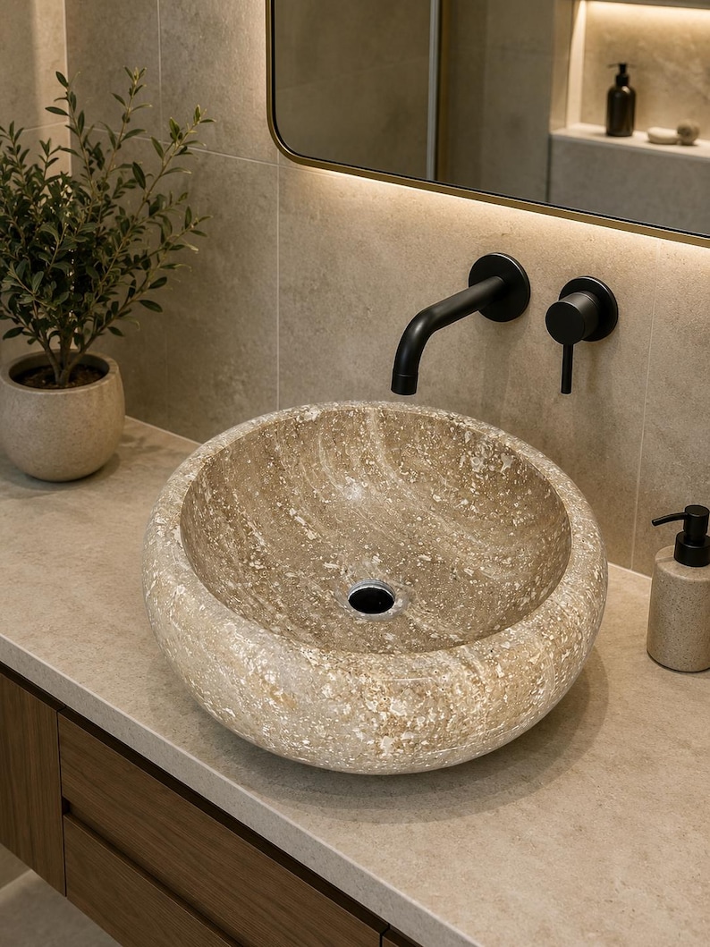 May include: A round, beige stone vessel sink with a textured, mottled surface and a central drain hole. The sink is mounted on a light-colored countertop and paired with a matte black wall-mounted faucet and a matching circular handle control.