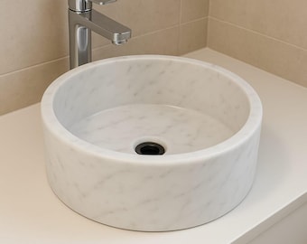 Op de afbeelding: Een ronde, witte marmeren wastafel met een zwarte afvoer. Een zilverkleurige kraan is aan de muur bevestigd. De wastafel staat op een wit aanrechtblad. Het ontwerp is modern en minimalistisch. De diameter is ongeveer 40 cm.