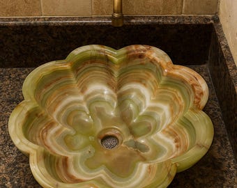 May include: A flower-shaped onyx sink with green, white, and brown veining. The sink is set into a dark granite countertop, with a bronze faucet and a tiled wall in the background. The sink has a drain in the center.