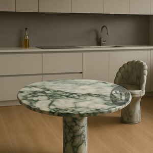 Puede incluir: Una mesa redonda de m&aacute;rmol con un patr&oacute;n veteado verde y blanco, sostenida por una base cil&iacute;ndrica. Una silla a juego con respaldo en forma de concha est&aacute; al lado de la mesa. La mesa est&aacute; en una cocina con armarios claros y suelo de madera.