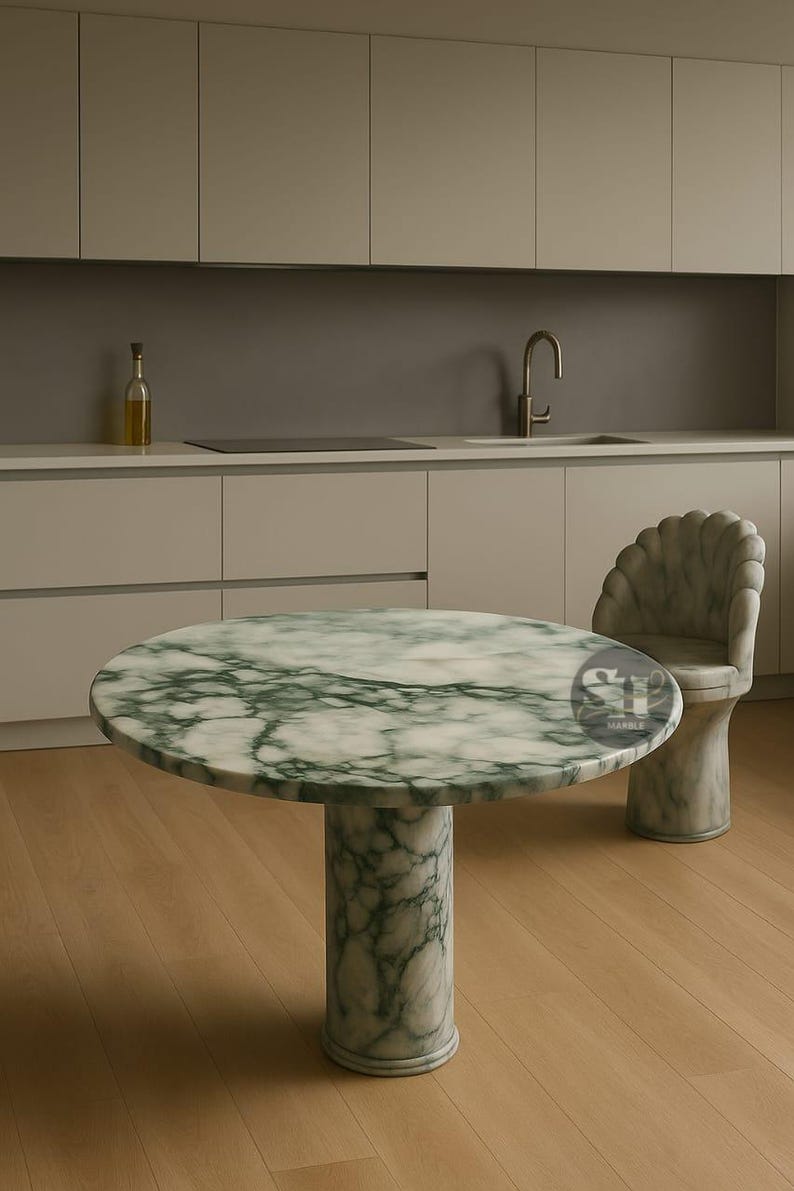 Puede incluir: Una mesa redonda de m&aacute;rmol con un patr&oacute;n veteado verde y blanco, sostenida por una base cil&iacute;ndrica. Una silla a juego con respaldo en forma de concha est&aacute; al lado de la mesa. La mesa est&aacute; en una cocina con armarios claros y suelo de madera.