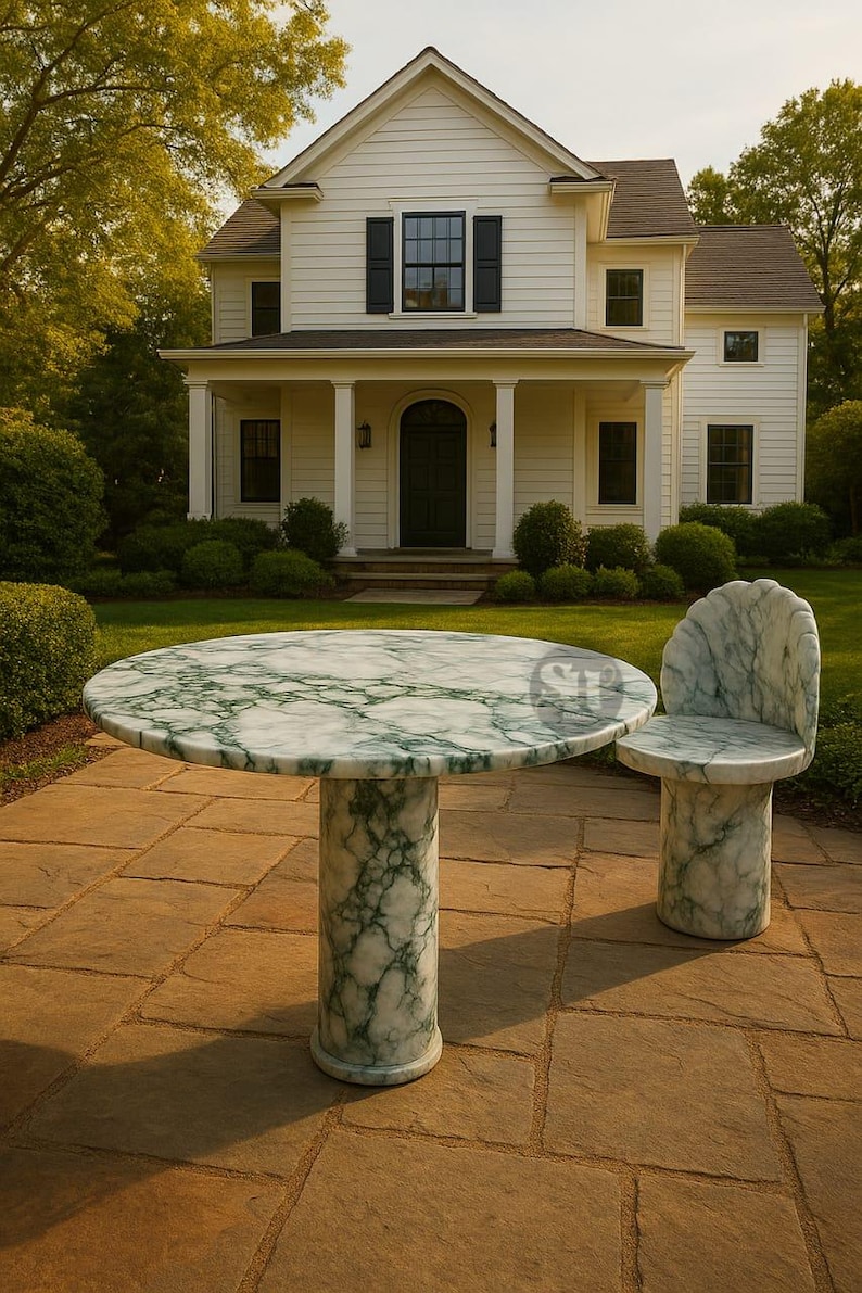 Puede incluir: Una mesa redonda de m&aacute;rmol y una silla a juego con vetas verdes. La mesa tiene una base de pedestal &uacute;nico. La silla tiene respaldo curvo. El conjunto est&aacute; al aire libre en un patio de piedra frente a una casa blanca.