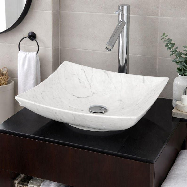 May include: A white marble square vessel sink with gray veining. The sink sits on a dark brown vanity with a chrome faucet. A white towel hangs on a black towel ring, and a small vase with greenery is visible.