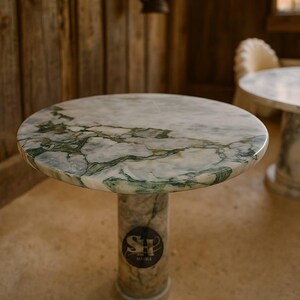 Puede incluir: Mesa redonda de m&aacute;rmol con tapa veteada blanca y verde. La mesa tiene una base cil&iacute;ndrica con un logotipo. La mesa est&aacute; en una habitaci&oacute;n con paredes de madera y suelo de hormig&oacute;n.