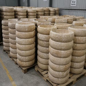 May include: Multiple stacks of round beige marble vessel sinks arranged on wooden pallets in a warehouse. Signs on the wall read Natural Stone and All Sink Stock Beige Marble. The stone basins feature a textured, mottled surface in shades of tan.