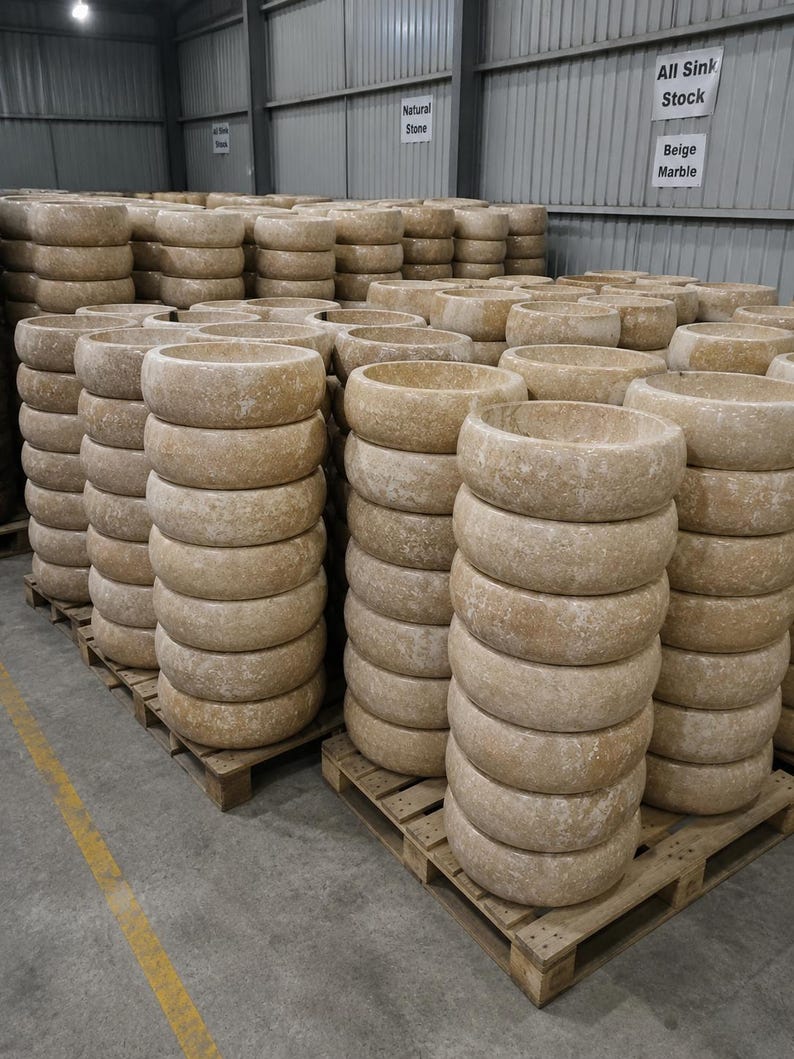 May include: Multiple stacks of round beige marble vessel sinks arranged on wooden pallets in a warehouse. Signs on the wall read Natural Stone and All Sink Stock Beige Marble. The stone basins feature a textured, mottled surface in shades of tan.