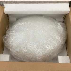 May include: A round object wrapped in clear plastic bubble wrap, securely positioned inside a brown cardboard shipping box. The item is cushioned by white rectangular foam inserts placed along the top and bottom edges of the interior container.