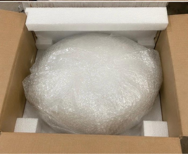 May include: A round object wrapped in clear plastic bubble wrap, securely positioned inside a brown cardboard shipping box. The item is cushioned by white rectangular foam inserts placed along the top and bottom edges of the interior container.