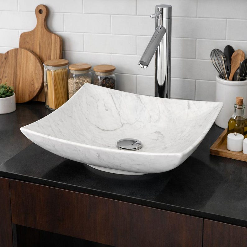 May include: A white marble square vessel sink with gray veining. The sink is set on a dark countertop with a chrome faucet. Kitchen accessories, including wooden cutting boards and jars, are in the background.