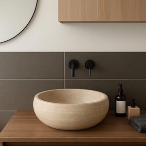 May include: A round, beige stone vessel sink sits on a wooden vanity. Behind the sink, two matte black wall-mounted faucets are installed against a dark gray tiled wall. A dark amber soap dispenser and a small wooden container are placed nearby.