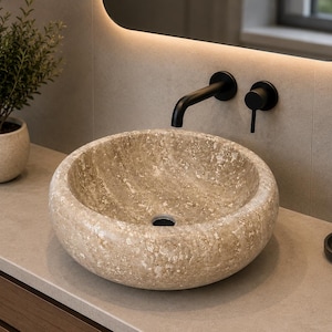 May include: A round vessel sink crafted from beige natural stone with a textured, mottled surface. The sink features a central drain hole and is paired with a matte black wall-mounted faucet and a matching black handle control fixture.