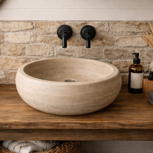 May include: A round, beige stone vessel sink with a smooth, natural texture and a central drain. The basin features subtle horizontal striations in the stone, creating a neutral appearance that sits atop a wooden vanity surface.