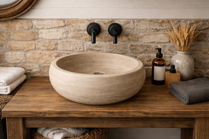 May include: A round, beige stone vessel sink with a smooth, natural texture and a central drain. The basin features subtle horizontal striations in the stone, creating a neutral appearance that sits atop a wooden vanity surface.