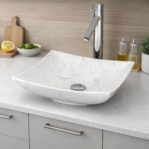 May include: A square, white marble vessel sink with a chrome faucet. The sink has a polished finish and sits on a gray countertop. A wooden cutting board, a bowl of limes, and a lemon are in the background.