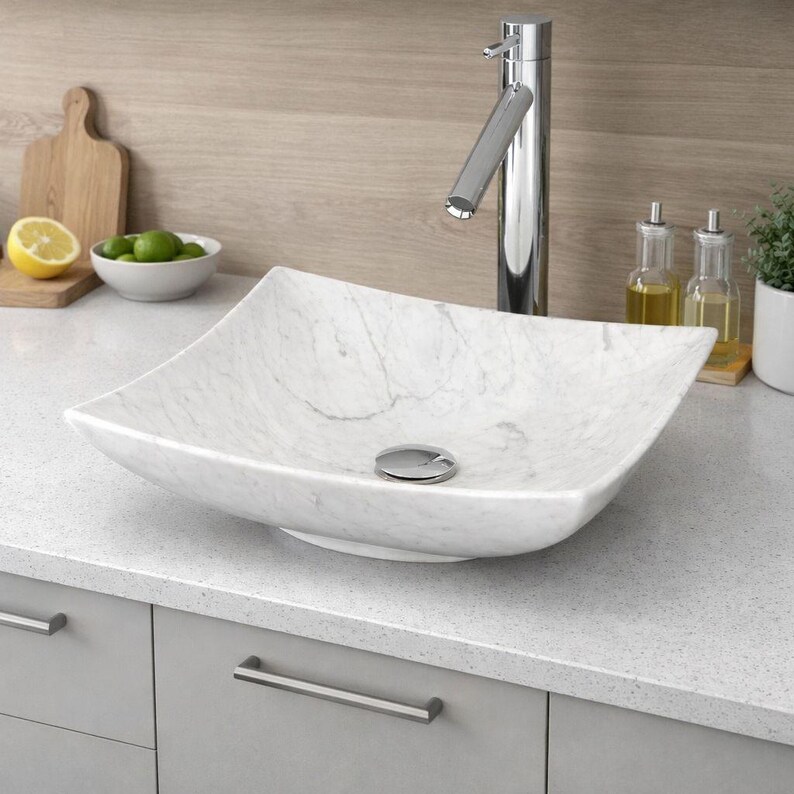 May include: A square, white marble vessel sink with a chrome faucet. The sink has a polished finish and sits on a gray countertop. A wooden cutting board, a bowl of limes, and a lemon are in the background.