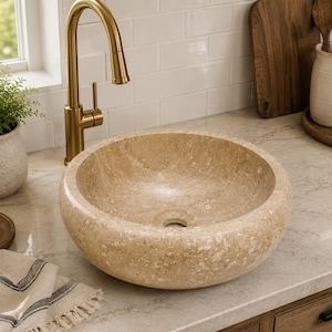 May include: A round vessel sink crafted from beige natural stone with a textured, porous surface and a central drain hole. The basin features a consistent circular shape with thick, rounded edges, showcasing the organic variations of the stone.