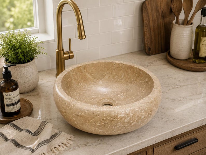 May include: A round vessel sink crafted from beige natural stone with a textured, porous surface and a central drain hole. The basin features a consistent circular shape with thick, rounded edges, showcasing the organic variations of the stone.