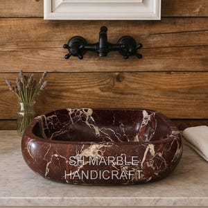 May include: A rectangular, reddish-brown marble sink with white veining, featuring the text "SH MARBLE HANDICRAFT". The sink is set on a light-colored countertop, with a black faucet and a wooden background.