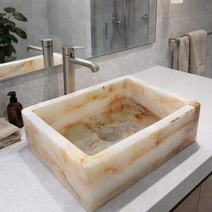 May include: Rectangular onyx bathroom sink with a natural, veined pattern in shades of cream, beige, and orange. The sink has a drain and is set on a white countertop. A stainless steel faucet is visible.