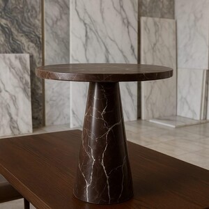 May include: A brown marble side table with a round top and a conical base. The table is set on a dark brown wooden surface. The marble has white veining. The table is approximately 20 inches in diameter and 24 inches tall.