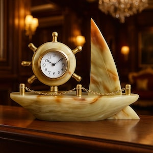 May include: A decorative clock in the shape of a sailboat. The clock face is round, with a gold-colored bezel and a white face. The sailboat is made of a light green and brown stone, with a gold-colored chain and accents. The clock measures approximately 8 inches long.