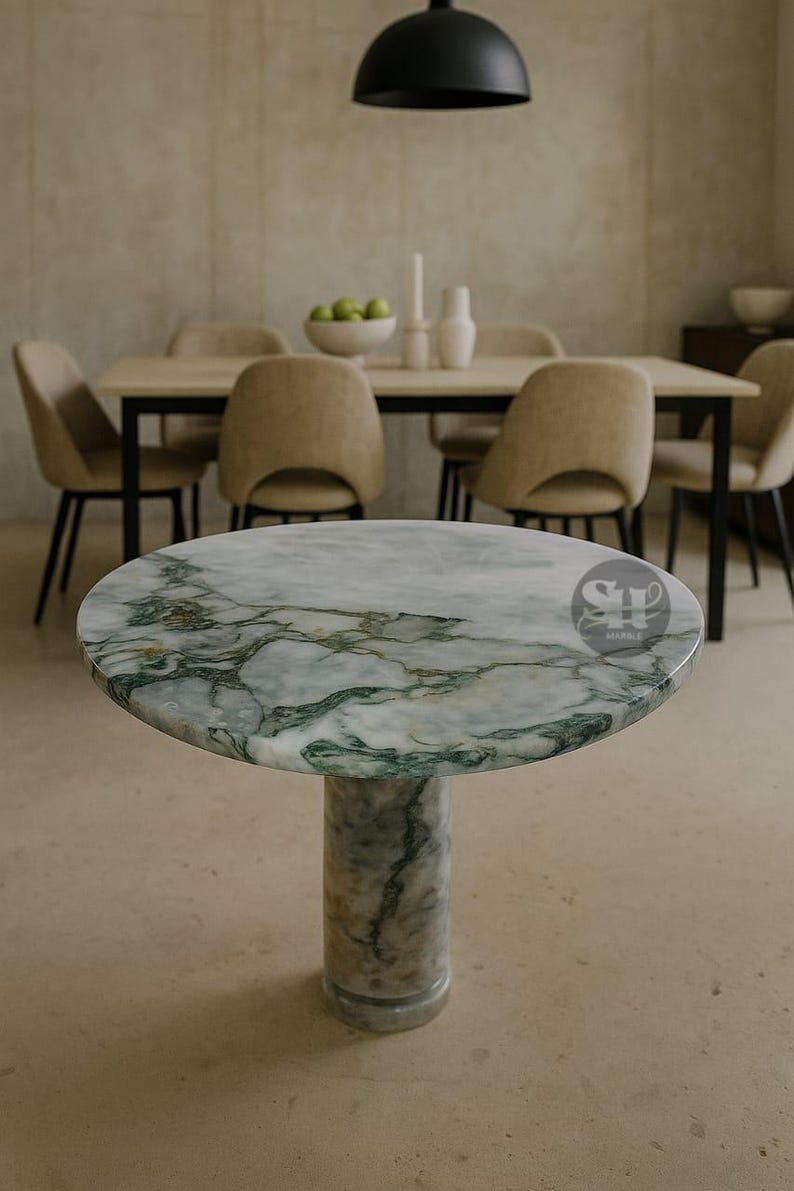 Puede incluir: Una mesa redonda de m&aacute;rmol con un patr&oacute;n veteado verde y blanco. La mesa tiene una base de pedestal cil&iacute;ndrica &uacute;nica. La mesa est&aacute; en un comedor con sillas y una mesa en el fondo.