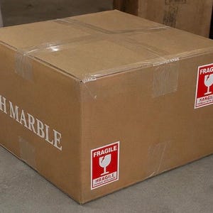 May include: A square brown cardboard shipping box sealed with clear tape. The side features the white text SH MARBLE. Two red and white rectangular stickers are attached, each displaying the word FRAGILE above a white wine glass icon.