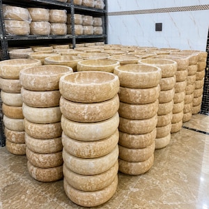 May include: A large collection of round, beige stone vessel sinks stacked in multiple columns. Each sink features a smooth, polished rim and a natural, mottled texture with variations in color, ranging from light cream to tan, characteristic of natural stone.