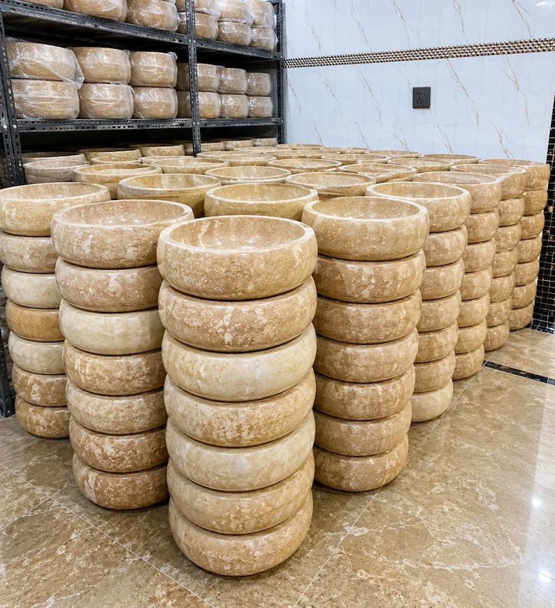 May include: A large collection of round, beige stone vessel sinks stacked in multiple columns. Each sink features a smooth, polished rim and a natural, mottled texture with variations in color, ranging from light cream to tan, characteristic of natural stone.