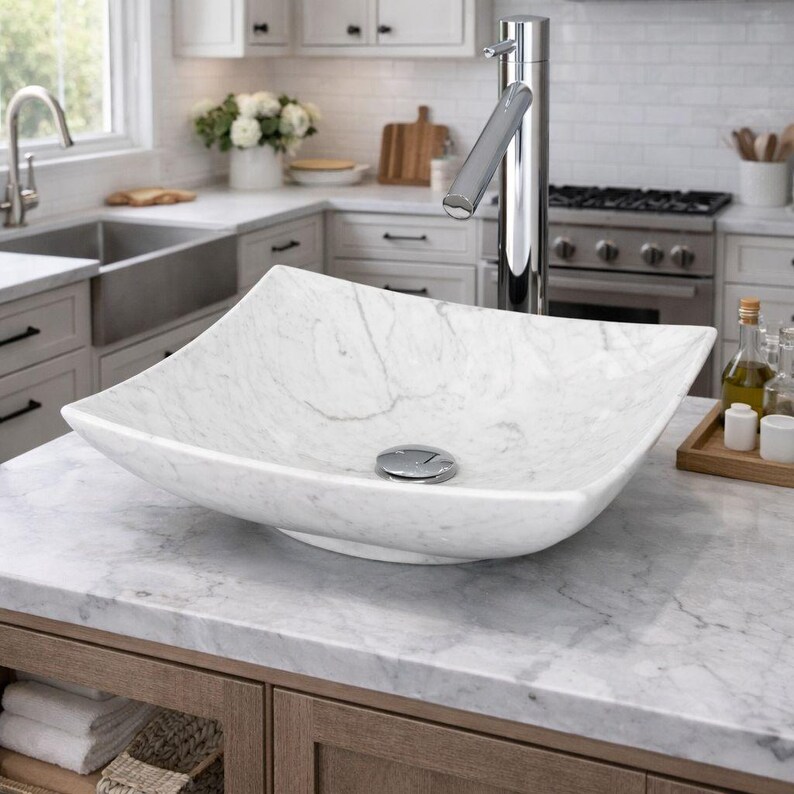 May include: A white marble vessel sink with a square shape and curved edges. The sink has a polished chrome drain and sits on a marble countertop. A tall chrome faucet is positioned behind the sink. The kitchen setting includes a stainless steel sink and white cabinets.