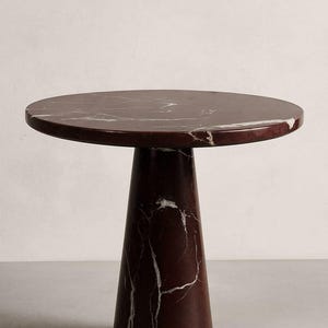 May include: A round side table with a dark red marble top and a matching conical base. The marble has white veining throughout. The table is a modern design, suitable for a living room or bedroom.