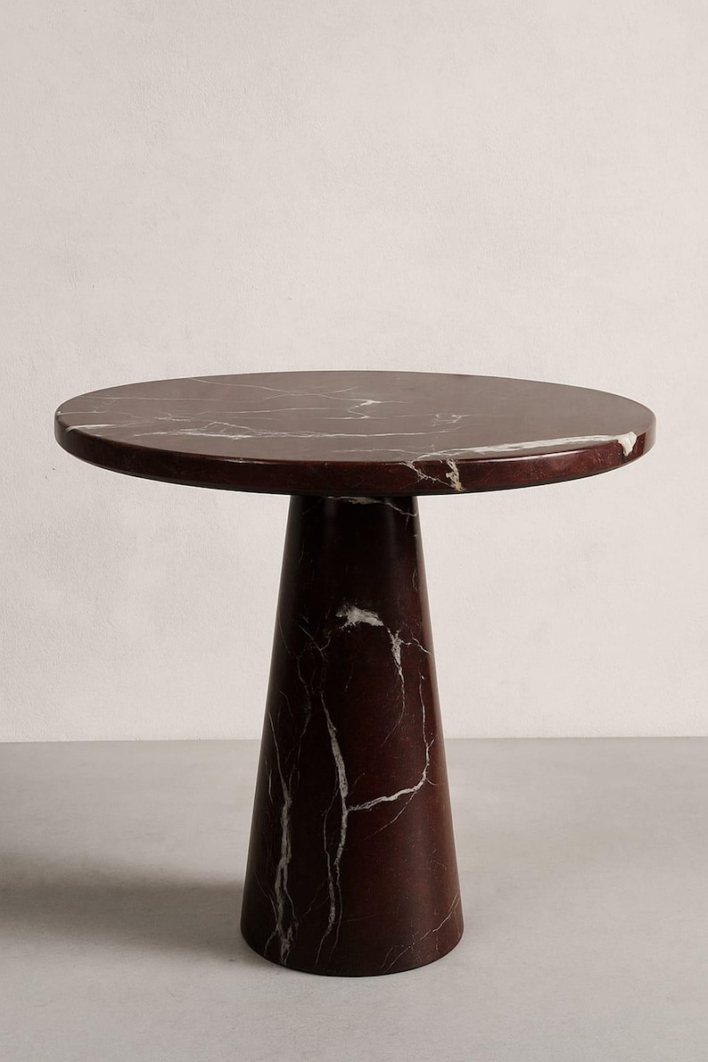May include: A round side table with a dark red marble top and a matching conical base. The marble has white veining throughout. The table is a modern design, suitable for a living room or bedroom.