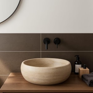 May include: A round, beige stone vessel sink with a natural textured finish sits on a wooden vanity. Above the sink, two matte black wall-mounted faucet fixtures are installed against a dark brown tiled wall, accompanied by a soap dispenser.
