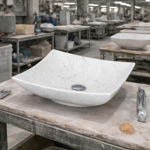 May include: A white marble square vessel sink with a polished chrome drain. The sink has a gently curved shape and is set on a weathered wooden surface. The background shows a workshop setting with other sinks.