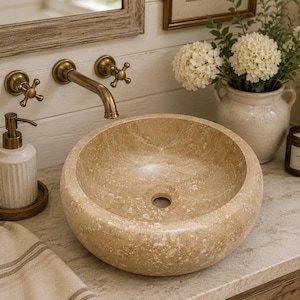May include: A round, beige stone vessel sink with a textured, natural finish and a central drain hole. The basin features a smooth interior surface and a porous, mottled exterior, showcasing the organic variations of the natural stone material.