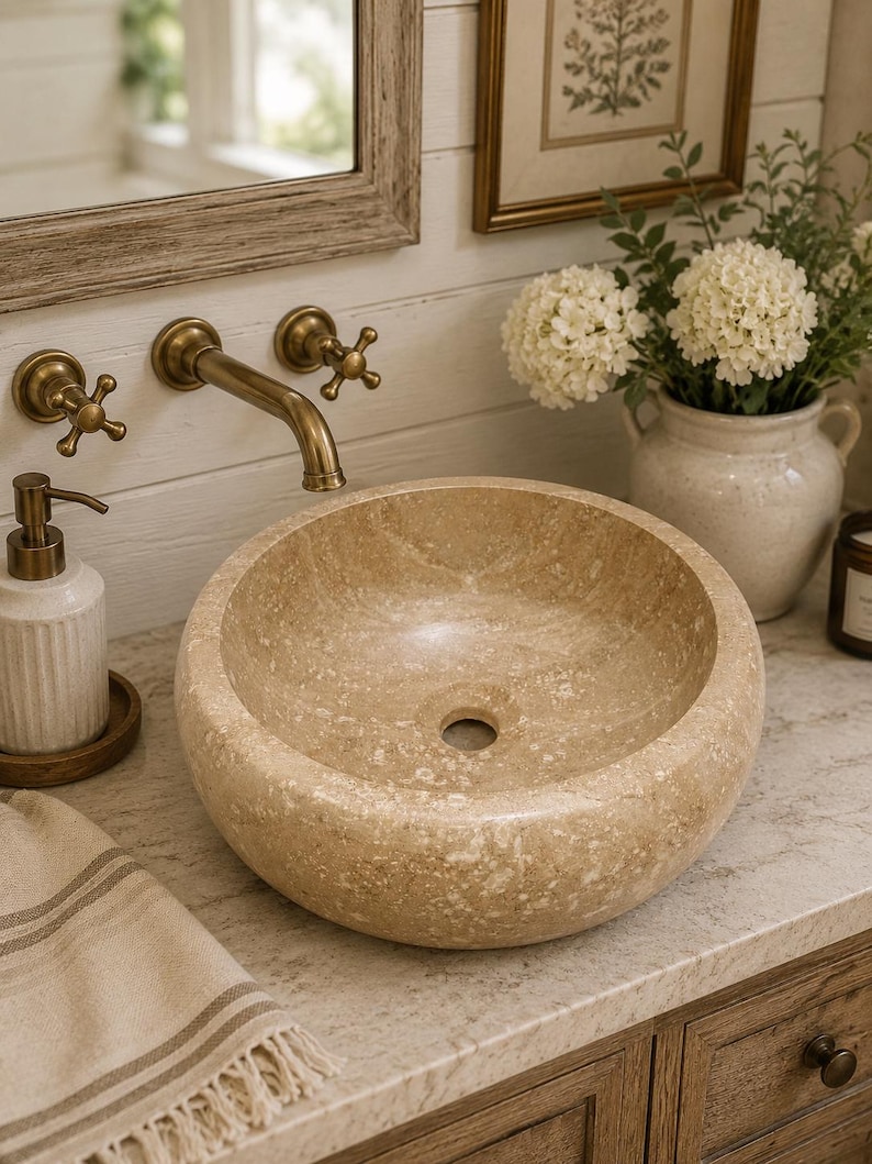 May include: A round, beige stone vessel sink with a textured, natural finish and a central drain hole. The basin features a smooth interior surface and a porous, mottled exterior, showcasing the organic variations of the natural stone material.