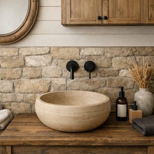 May include: A round, beige stone vessel sink sits on a dark wooden vanity. Above the sink, two matte black wall-mounted faucets are installed into a light-colored stone wall. A round mirror and wooden cabinets are mounted on the wall above.
