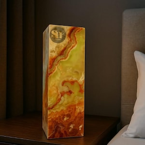 May include: A rectangular onyx lamp with a warm color palette of yellow, orange, and red. The lamp is placed on a dark wooden nightstand next to a bed with a white pillow. The lamp has a logo on the top.