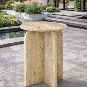 May include: A round side table crafted from beige travertine stone, featuring a textured, porous surface with natural tan and cream veining. The table is supported by a central base composed of three vertical panels arranged in a cross shape.