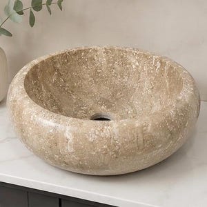 May include: A round vessel sink crafted from beige natural stone with a polished finish and visible mineral flecks. The basin features a smooth, curved interior and a central drain hole, showcasing the unique texture and color variations of the stone.