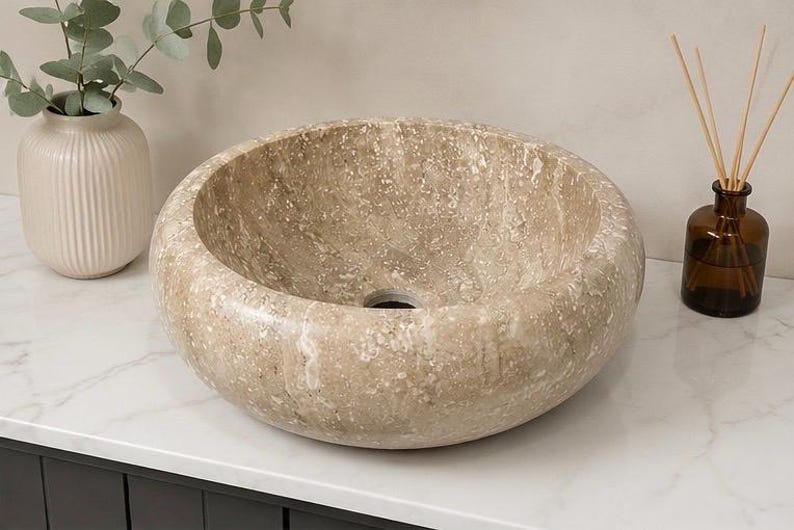 May include: A round vessel sink crafted from beige natural stone with a polished finish and visible mineral flecks. The basin features a smooth, curved interior and a central drain hole, showcasing the unique texture and color variations of the stone.
