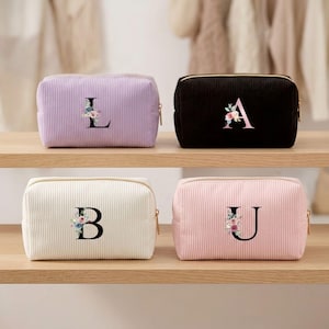 Könnte beinhalten: Vier rechteckige Kosmetiktaschen aus Kord in Lavendel, Schwarz, Creme und Hellrosa. Jede Tasche zeigt einen großen, zentrierten Serifenbuchstaben—L, A, B oder U—verziert mit einem kleinen, bunten Blumenarrangement im Aquarellstil und einem goldenen Reißverschluss.