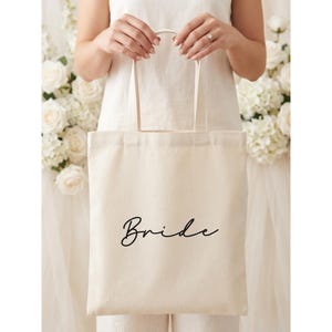 Könnte beinhalten: Eine cremefarbene Stofftasche aus Segeltuch mit dem Wort Bride in schwarzer Schreibschrift. Die Tasche hat eine rechteckige Form mit zwei passenden Stoffhenkeln an der Oberseite und besteht aus einem schlichten Webstoff.
