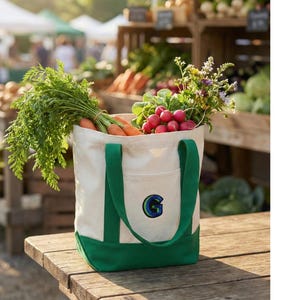 Könnte beinhalten: Eine cremefarbene Canvas-Tragetasche mit grünen Griffen und Boden, gefüllt mit frischem Gemüse und Blumen. Die Tasche enthält Karotten, Radieschen und Blumen. Auf der Tasche ist ein blaues, gesticktes "G". Die Tasche steht auf einem Holztisch.