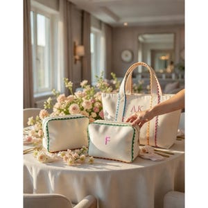 Könnte beinhalten: Off-white Canvas-Tasche und Kosmetiktaschen mit farbigem Besatz und Initialen-Stickerei. Die Tragetasche hat einen gewellten Rand und passende Griffe. Die Kosmetiktaschen haben grüne Verzierungen und einen Reißverschluss. Die Taschen liegen auf einer weißen Tischdecke.